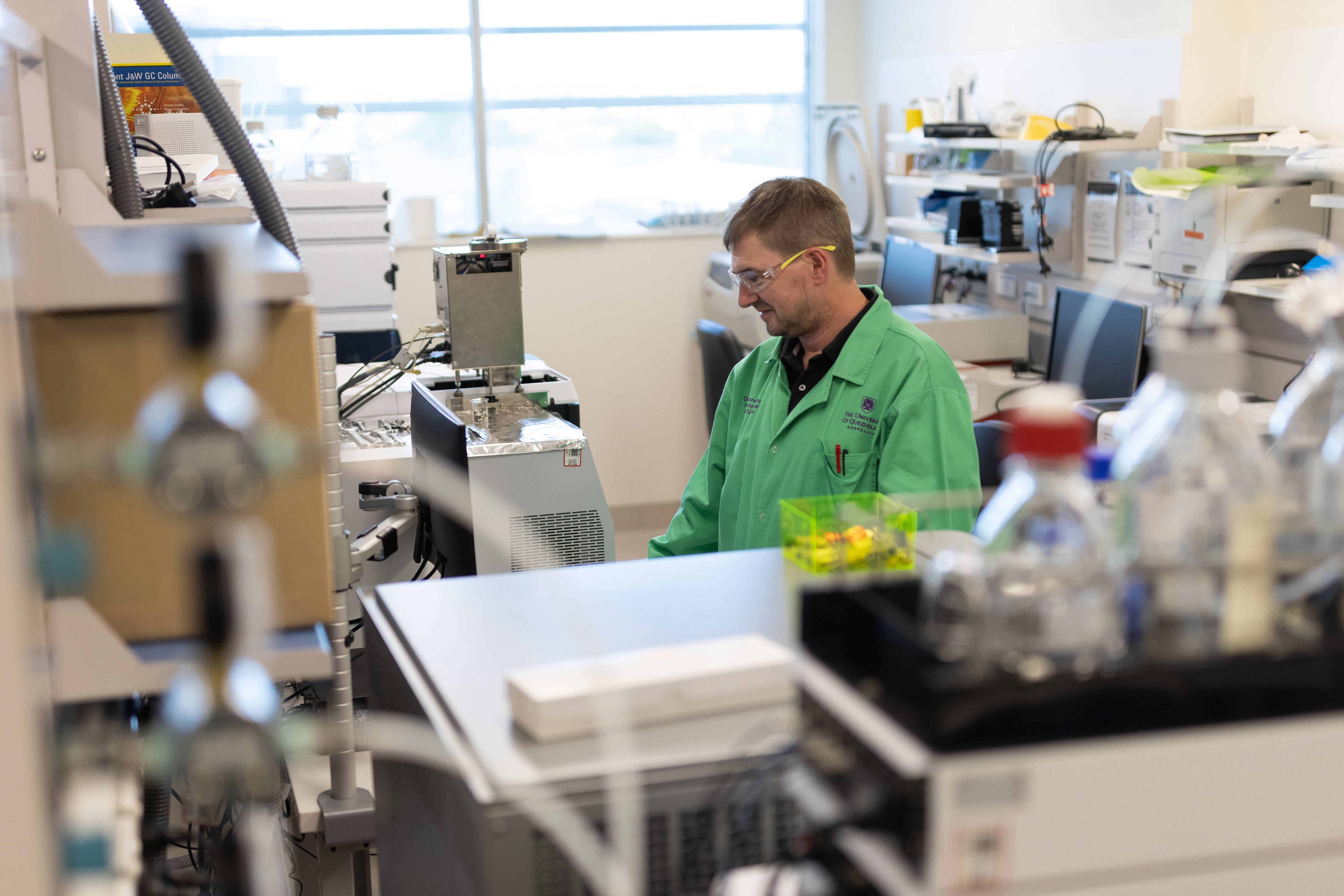 Prof Kevin Thomas in a QAEHS laboratory setting. Working on a laboratory instrument, wearing a green lab coat and protective eyewear. Photo: The University of Queensland.
