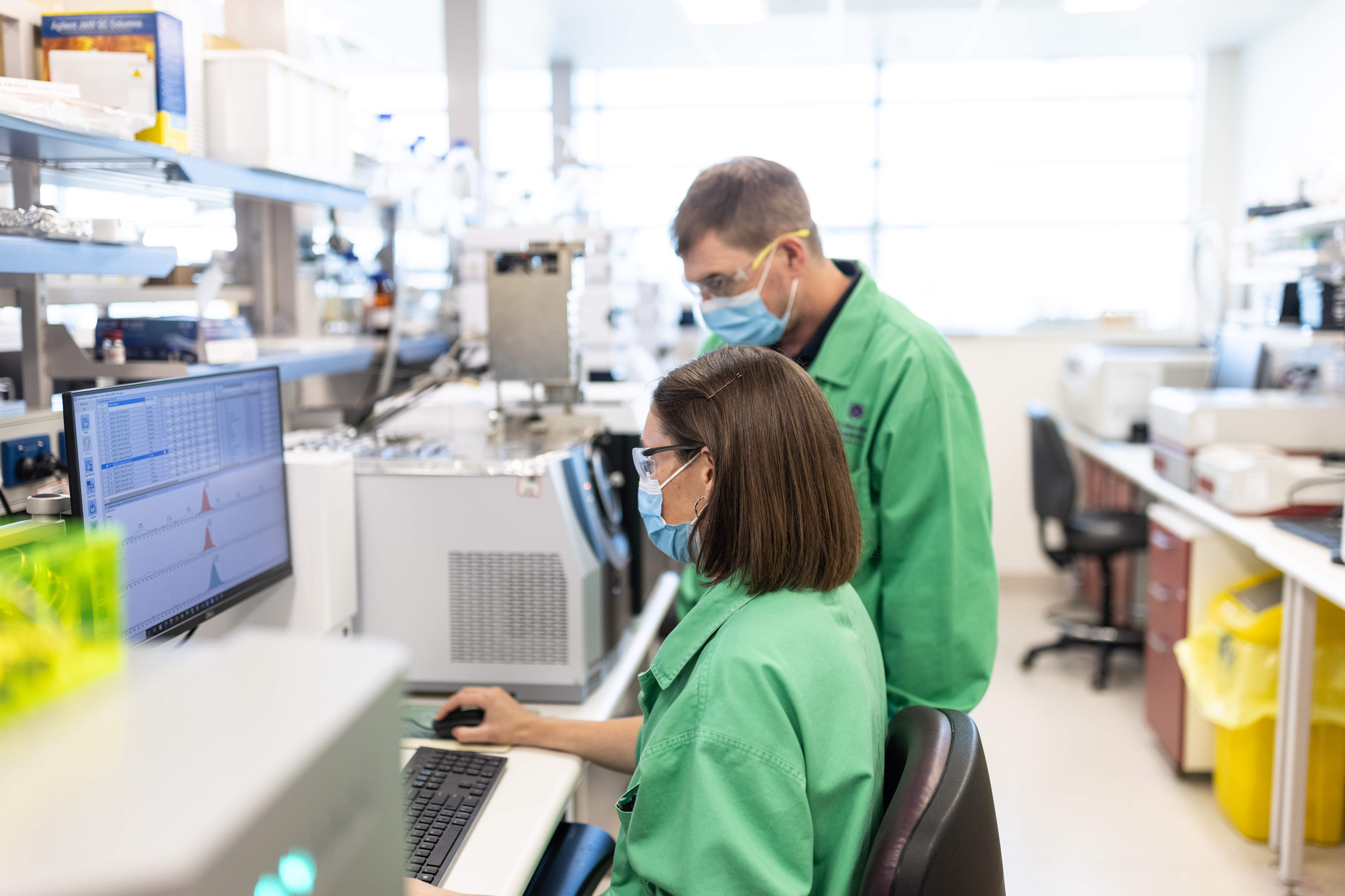 Prof Kevin Thomas in a QAEHS laboratory setting with Dr Cassie Rauert. Both looking at experimental results on a computer screen, wearing green lab coats, masks and protective eyewear. Photo: The University of Queensland.