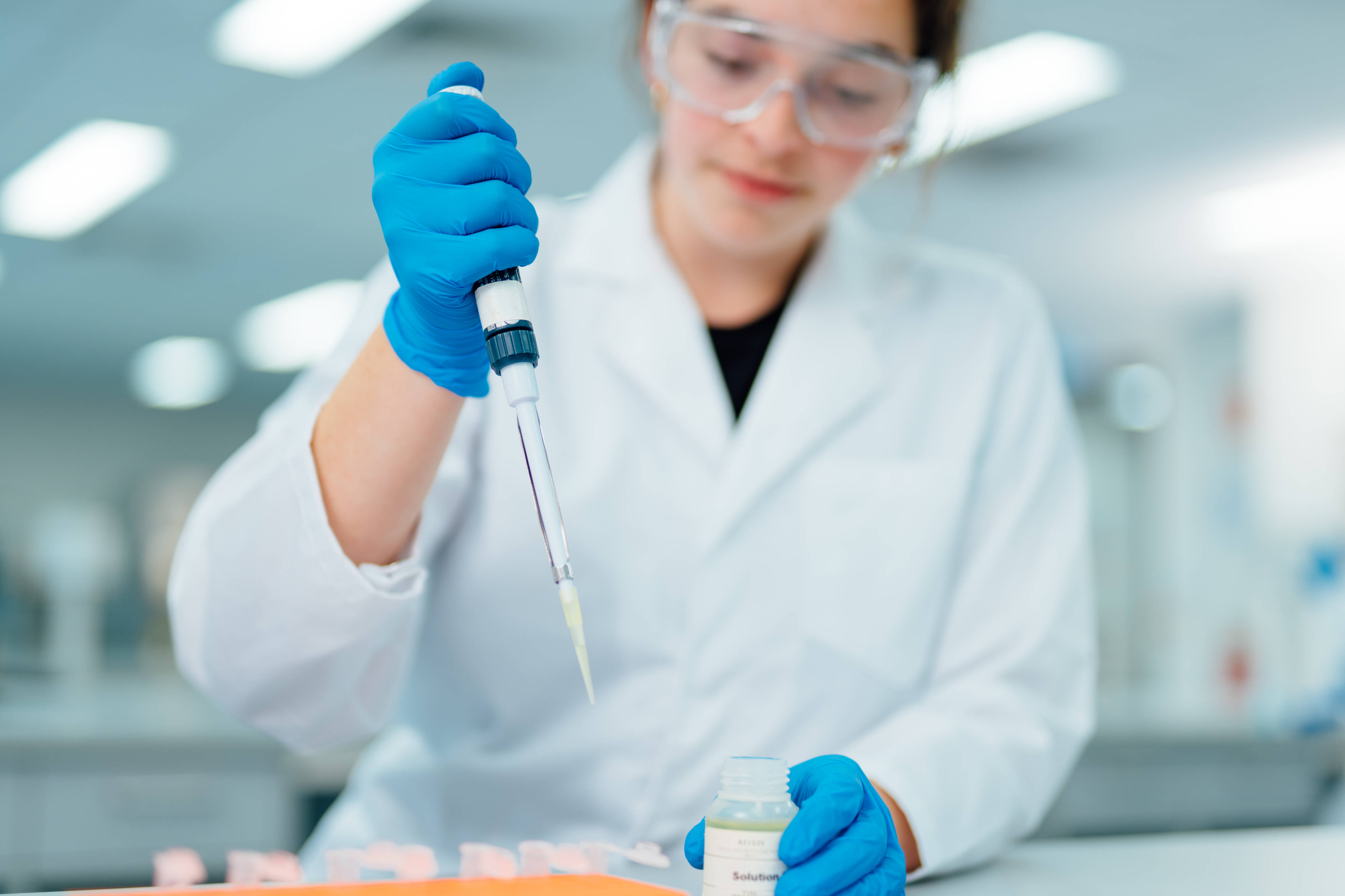 Young female scientist pipetting solution in a laboratory setting. Wearing eye protection, a white lab coat and blue gloves. Photo: University of Tasmania.