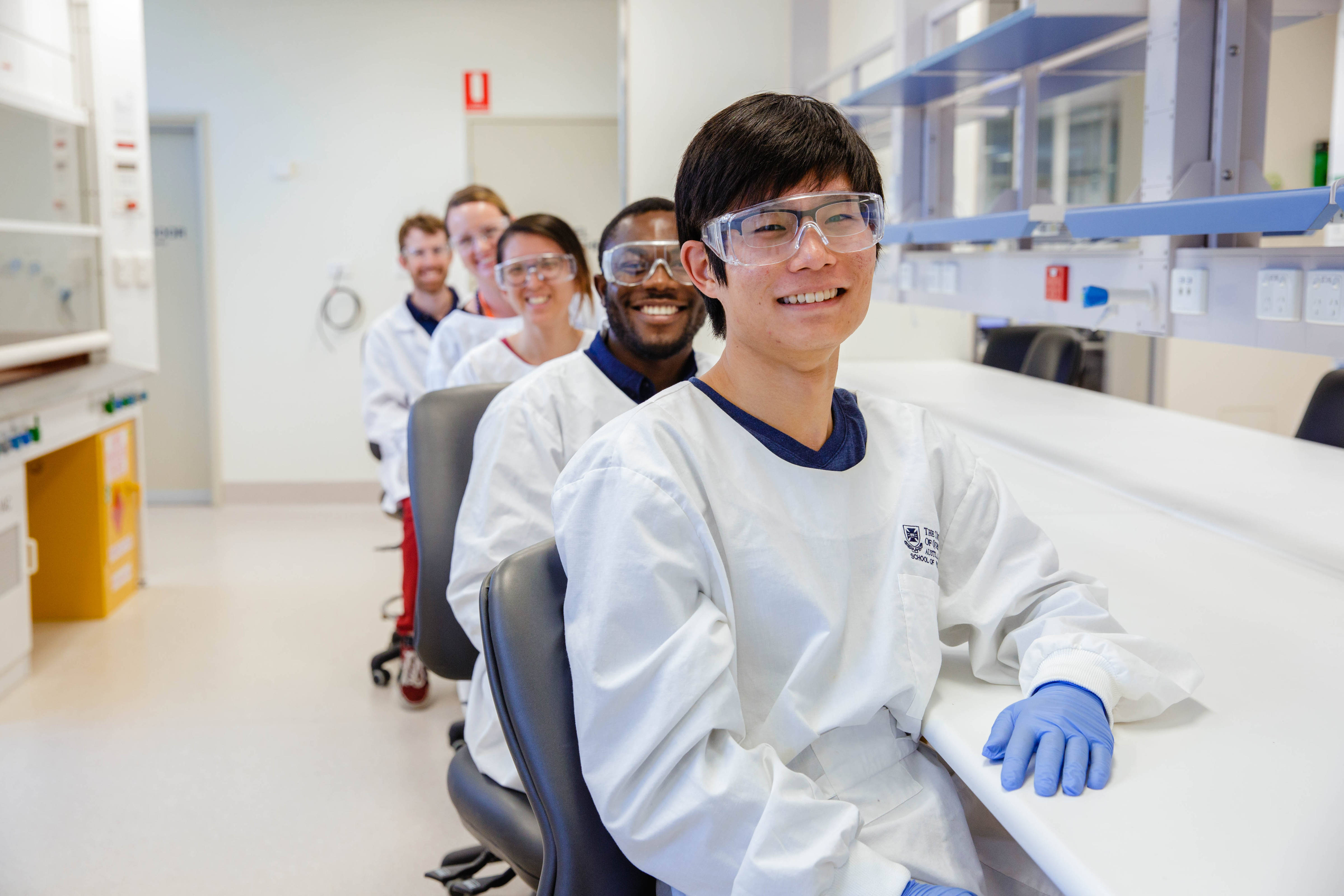 Five scientists dressed in PPE sitting at a QAEHS laboratory bench smiling with their chairs turned towards the camera. Photo: The University of Queensland.